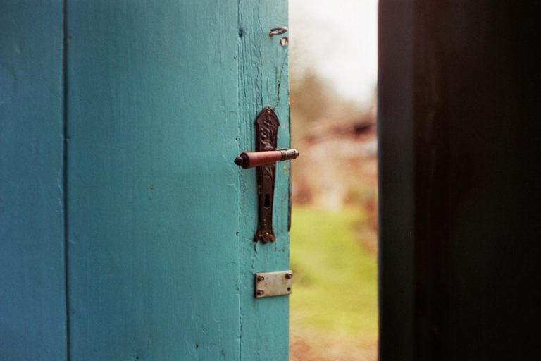A blue door with a metal handle that is slightly open revealing an out of focus green outdoor area beyond the door